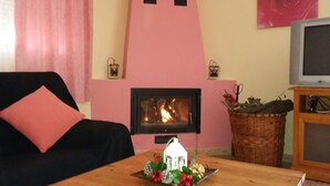 Een gezellige woonkamer met een open haard, een zwarte bank met een roze kussen, een klein tafeltje met een decoratie, en een televisie op een houten dressoir.