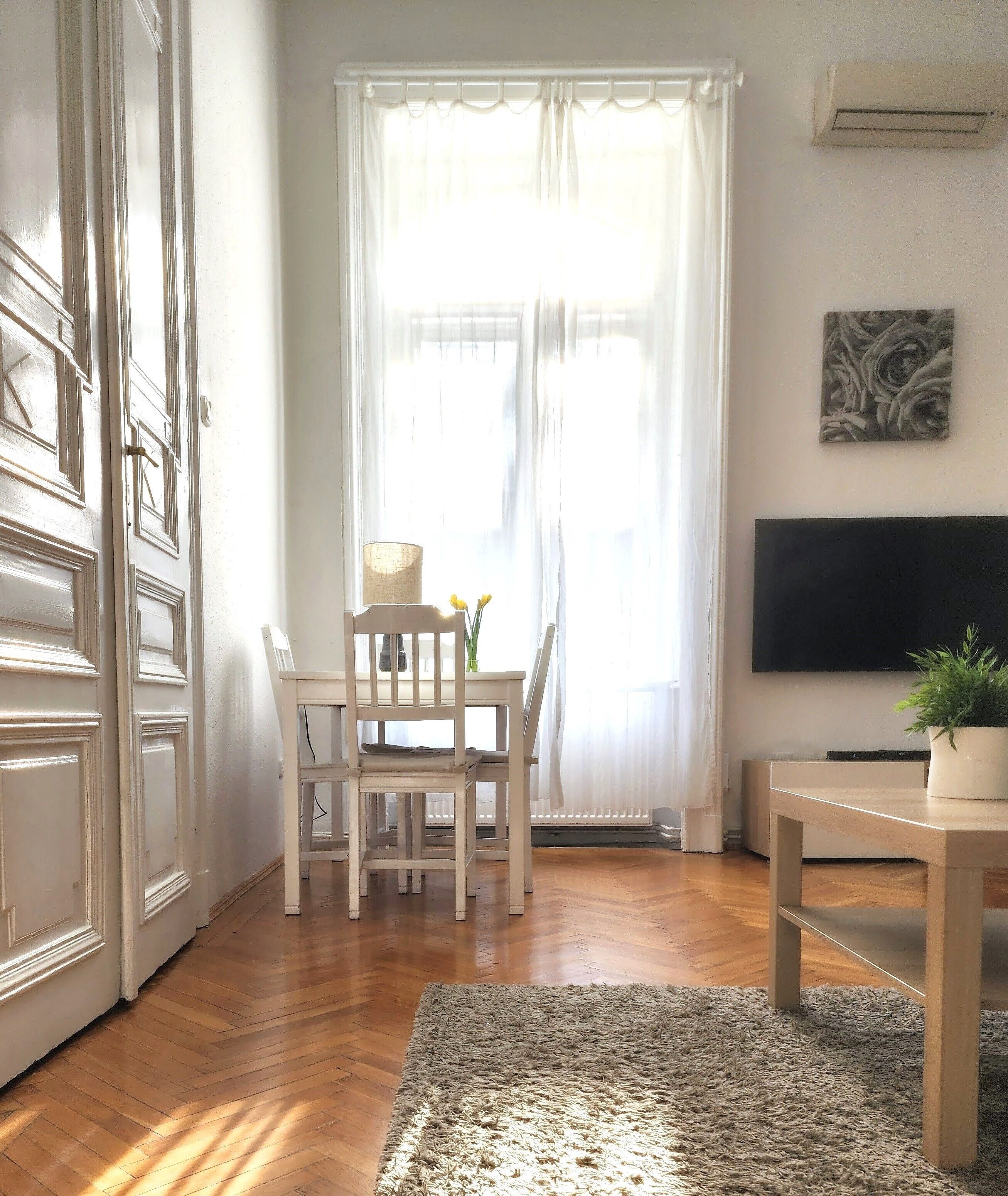 Et værelse med trægulv, et hvidt spisebord med stole, et lille bord med en plante, et fladskærms-tv på væggen og et vindue med let gardin.