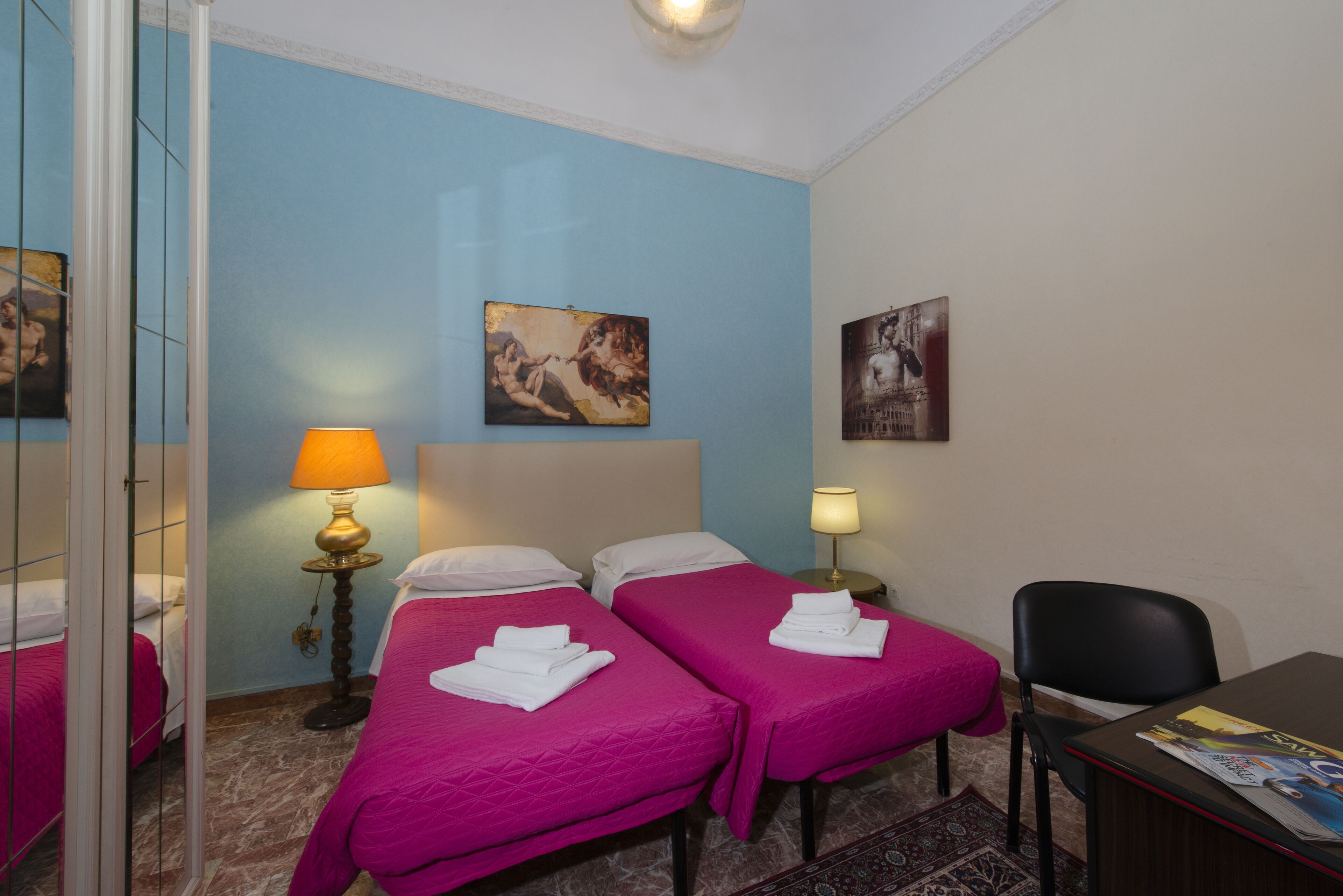 A hotel room with two beds, a desk, a chair, a lamp, and artwork on the walls.