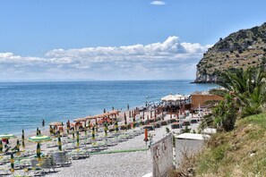 Uma praia com fileiras de espreguiçadeiras e guarda-sóis, um penhasco rochoso e o mar ao fundo.