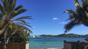 Una platja amb una tanca de fusta, palmeres i vista al mar amb vaixells.