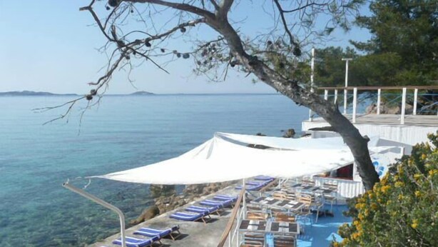 Zona junto a la piscina con sillas de descanso y sombrillas, con vista a un mar azul y transparente.