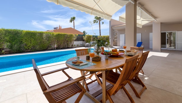 An outdoor dining area with a pool view, wooden chairs, a table with fruits and drinks, and a covered patio.