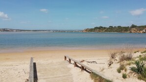 A beach with a wooden walkway leading to the water, sandy dunes, and a distant shoreline with buildings.