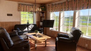 Sala de estar com sofá de couro preto, mesa de centro de madeira, televisão de tela plana e grandes janelas com vista para uma paisagem verde.