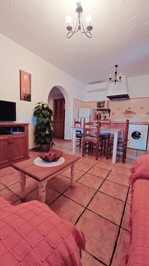 Uma sala de estar com televisão, uma mesa de jantar e cadeiras. Há uma planta e um lustre.