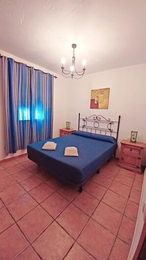 Un dormitorio con cama, candelabro, cortinas, mesita de noche y un cuadro en la pared.