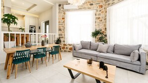 A modern living room with a grey sofa, wooden coffee table, and teal chairs. A kitchen with white cabinets and a stone accent wall is visible in the background. - Eumelia Iconic Villa, with Heated Pool & Whirlpool, BBQ, Close to amenities (Rethymno)