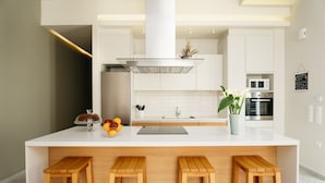A modern kitchen with white cabinetry, a central island with stools, a stainless steel range hood, and a built-in oven. - Eumelia Iconic Villa, with Heated Pool & Whirlpool, BBQ, Close to amenities (Rethymno)