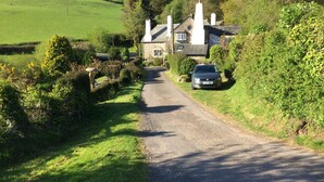 Eine Landstraße, die zu einem Steinhaus führt, vor dem ein Auto geparkt ist.