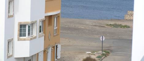 A coastal road with a parked car, buildings with balconies, and a view of the sea.