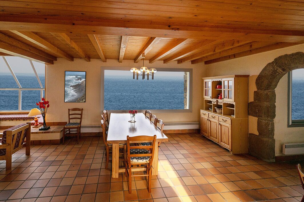 Un vaste espace dédié à la salle à manger, avec une grande table et des chaises, une vue sur l’océan par une grande fenêtre, et une cuisine dotée de meubles en bois.