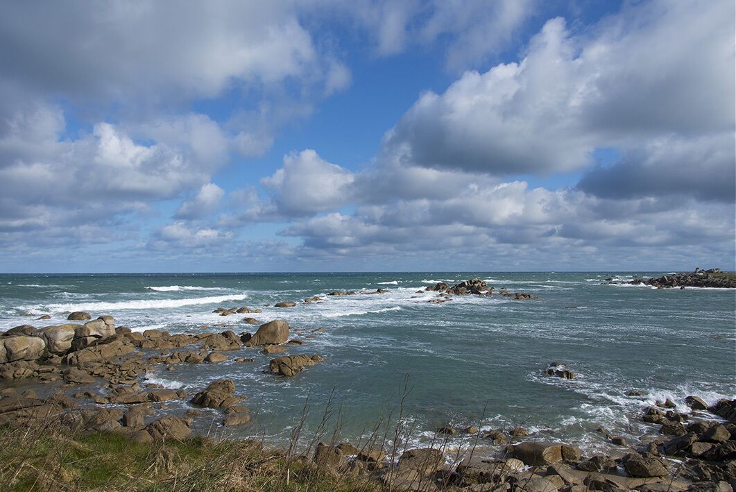 Une côte rocheuse où les vagues s’écrasent contre les rochers, sous un ciel partiellement nuageux.