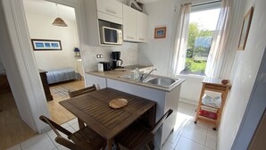 Une cuisine compacte équipée d’un four à micro-ondes, d’une machine à café et d’un évier. On y trouve une table à manger avec des chaises et une petite étagère sur laquelle est posé un bol. On aperçoit également un lit et une fenêtre avec des rideaux au fond de la pièce.