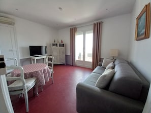 Een kamer met een grijze bank, een eettafel met een tafelkleed met stippen, een televisie op een standaard en een kast met decoratieve objecten.