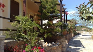 Un muro de piedra con macetas y flores, una pérgola de madera y un edificio con ventanas de arco.