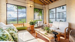 Una habitación iluminada por el sol, con ventanas amplias, un banco de madera, sofás blancos y diversas plantas en macetas.