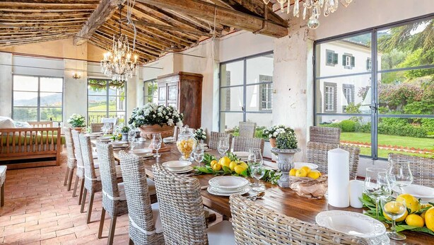 Un comedor con una larga mesa de madera dispuesta para una comida, rodeado de sillas de mimbre, con vista a zonas verdes y un edificio a través de amplios ventanales.