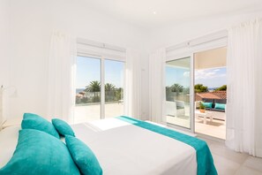 Quarto com cama grande, cortinas brancas e varanda com vista para palmeiras e um edifício.