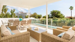Un espace extérieur aménagé avec des meubles en osier, une balustrade en verre et une vue sur une piscine et des bâtiments en arrière-plan, le tout baigné de lumière.