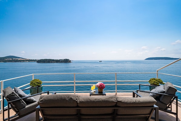 A deck with outdoor seating, a small table with a drink and flowers, and a view of the sea and distant islands.