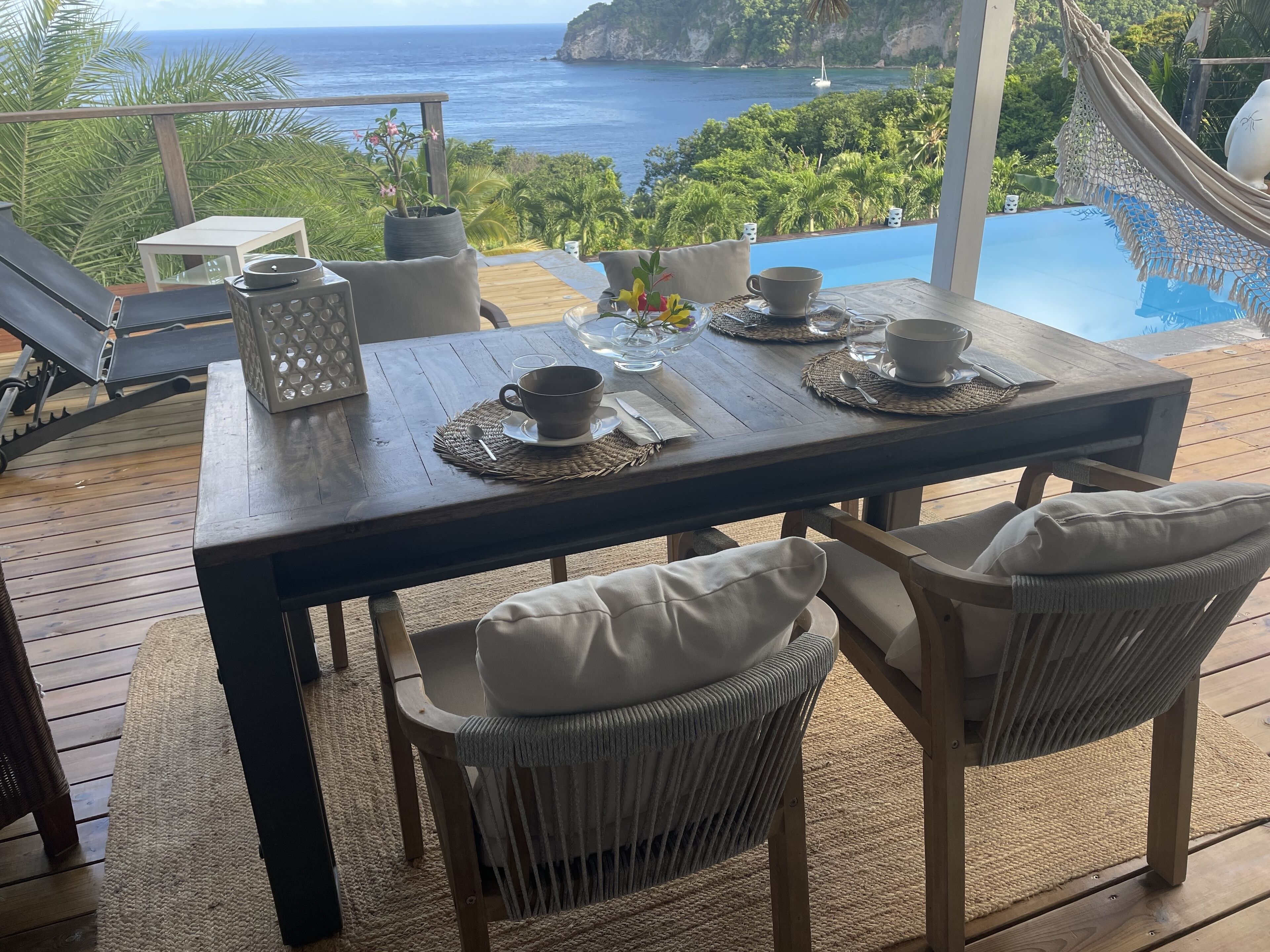A wooden deck with a table set for two, overlooking a pool and the ocean.