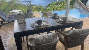 A wooden deck with a table set for two, overlooking a pool and the ocean. - SEA VIEW VILLA + OVERFLOW POOL (Deshaies)