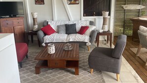 A cozy living room with a sofa, coffee table, and armchairs. There is a TV on a wooden cabinet, a painting on the wall, and a window with a view of greenery. - SEA VIEW VILLA + OVERFLOW POOL (Deshaies)