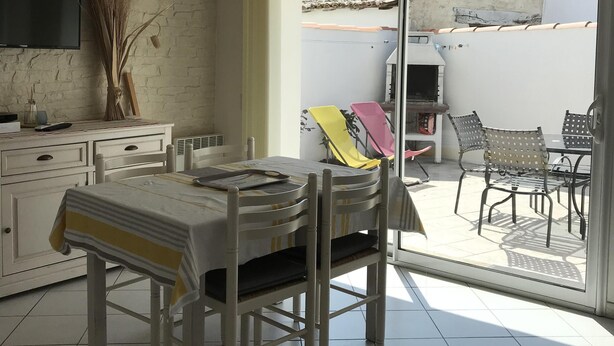 Un área de comedor con una mesa y sillas, un televisor instalado en una pared de ladrillo, y vistas del patio exterior con sillas y una mesa.