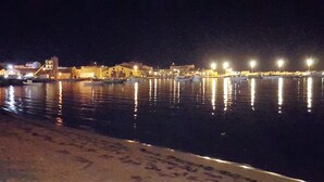 A nighttime waterfront scene with buildings and boats along the shore. - Villa with swimming pool facing the sea, for those who love peace and privacy (Marzamemi (Pachino) -SR)