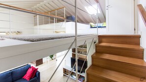 Un interior modern amb una escala de fusta, un altell amb barana de vidre i un sofà blau amb coixins vermells.
