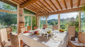 Un espace repas abrité, aménagé avec des meubles en osier, une table dressée avec des fruits et des verres, et une vue sur la campagne.
