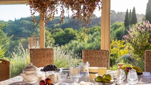 Une table dressée pour un repas, avec divers fruits, fromages et verres, donnant sur un jardin luxuriant.