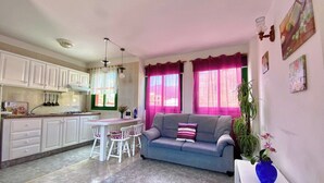 A compact kitchen with white cabinets, a dining area with a white table and chairs, a grey sofa with a purple pillow, and a blue vase on a white table.