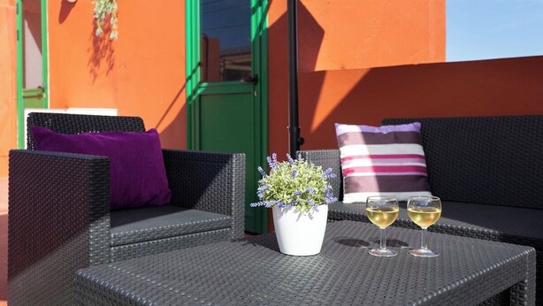 A wicker sofa with a purple pillow, a white pot with lavender, and two glasses of white wine on a table.