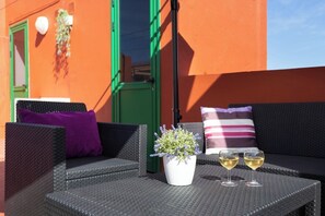 Un canapé en osier agrémenté d’un coussin violet, un pot blanc contenant de la lavande et deux verres de vin blanc posés sur une table.