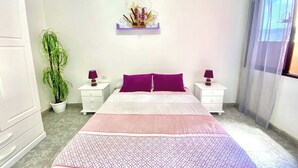 A bedroom with a bed, two nightstands, a potted plant, a wall-mounted shelf with decorative items, and a window with curtains.