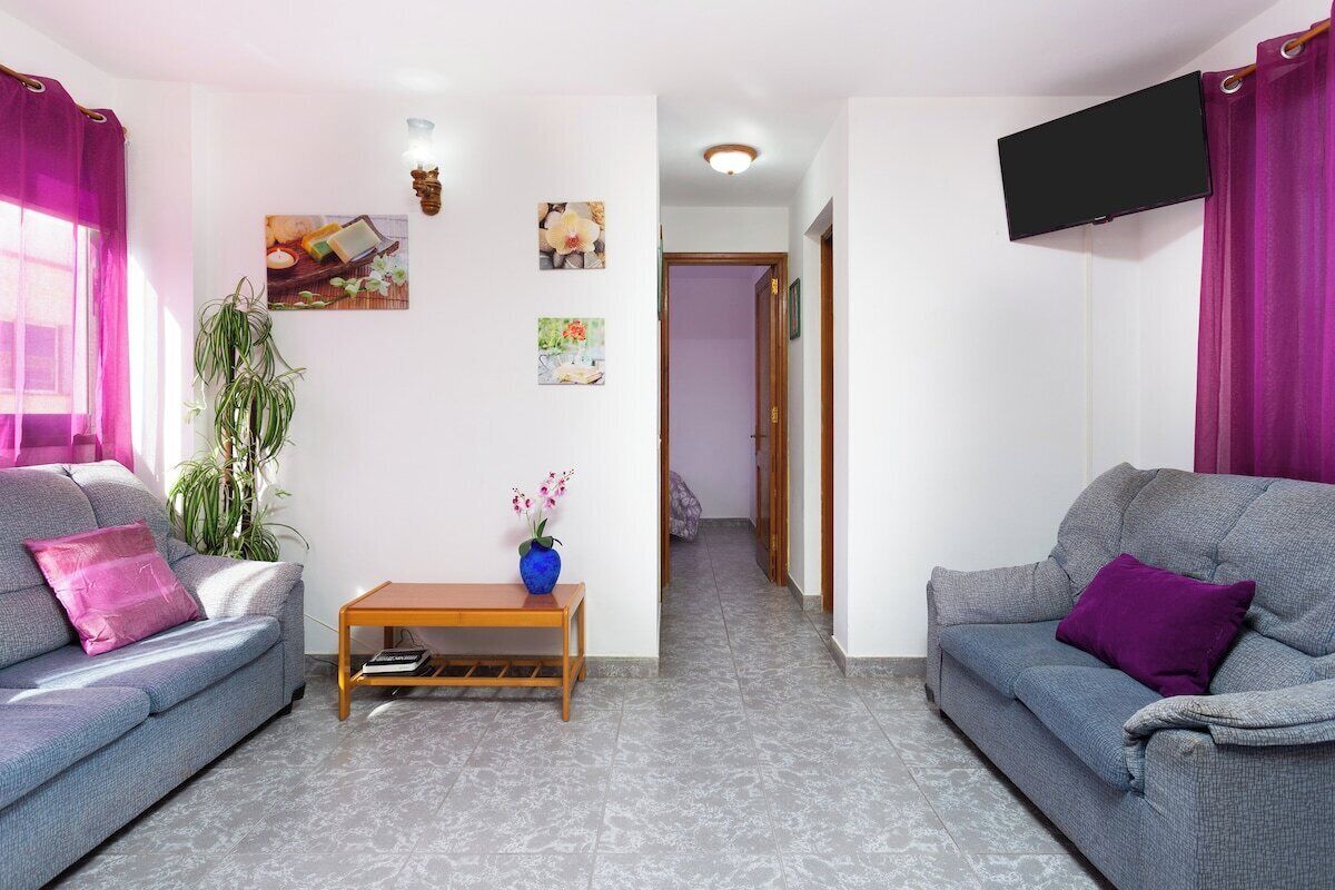 A living room with two grey sofas, a wooden coffee table, a potted plant, and a TV mounted on the wall.