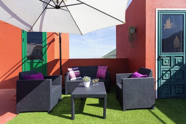 An outdoor seating area with wicker furniture, a table with a potted plant and glasses, and a white umbrella.