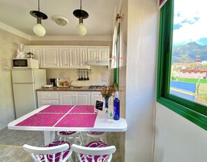 Une cuisine avec des meubles blancs, une table et des chaises roses, une fenêtre donnant sur des bâtiments et des montagnes, et un luminaire suspendu.