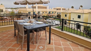 Une terrasse avec une table dressée pour le repas, un grand parasol et une vue sur les bâtiments environnants.