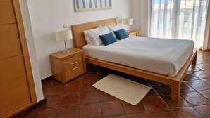 Une chambre à coucher avec un lit en bois, une literie blanche, deux lampes de chevet, une table de chevet avec un tiroir, un tableau au mur et une vue par une fenêtre avec des rideaux.