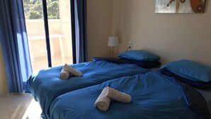 Habitación de hotel con dos camas, cada una con ropa de cama azul y toallas enrolladas. Se ve una ventana con cortinas azules, una lámpara en una mesita de noche y un cuadro en la pared.