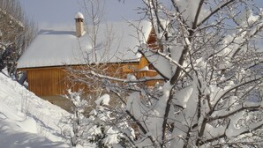 Une cabane en bois, dont le toit et la cheminée sont recouverts de neige, entourée d’arbres chargés de neige.