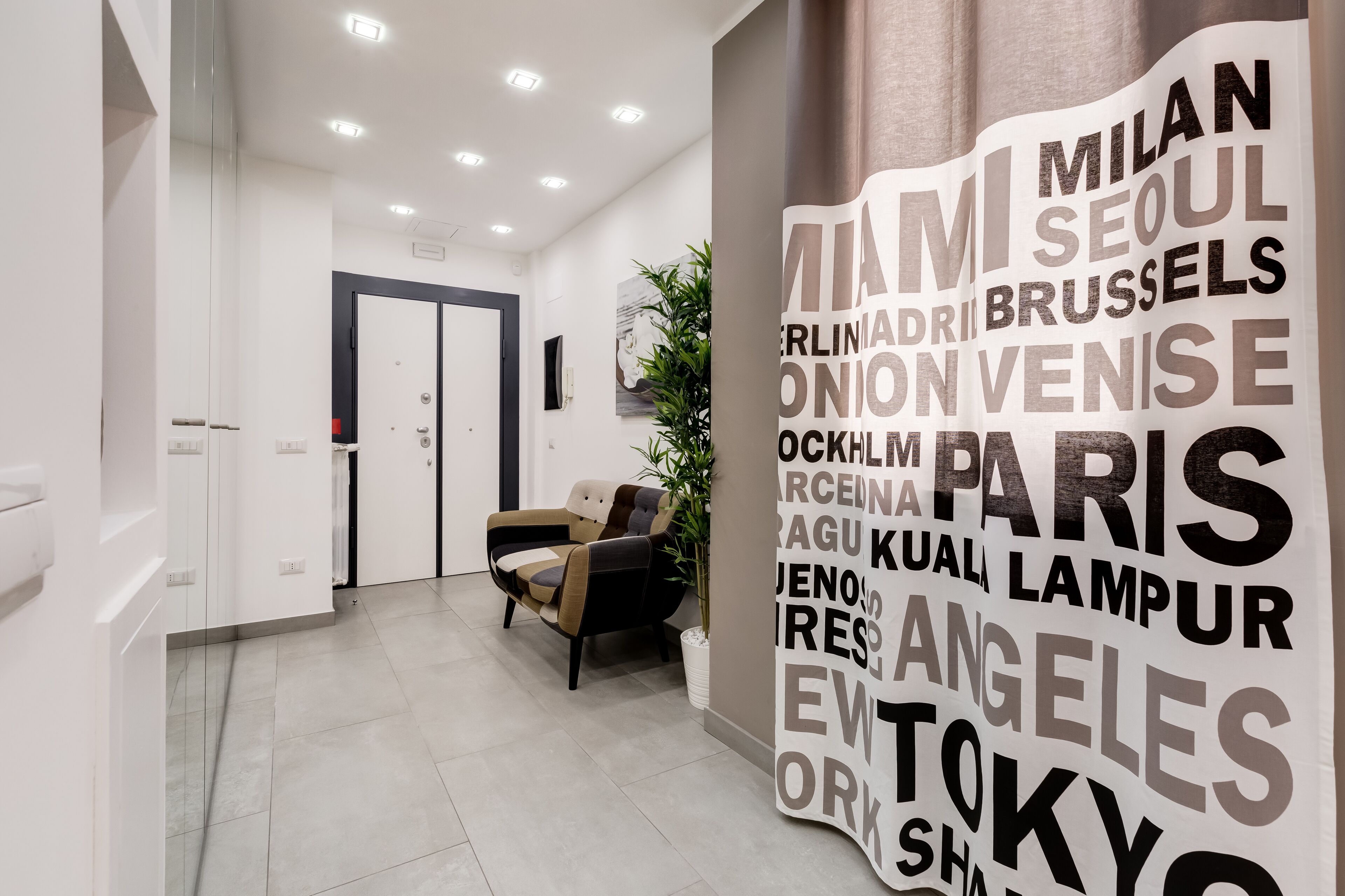 Un hall d’hôtel moderne comprenant un comptoir d’accueil, un espace salon avec un fauteuil et un mur sur lequel sont inscrits des noms de villes.