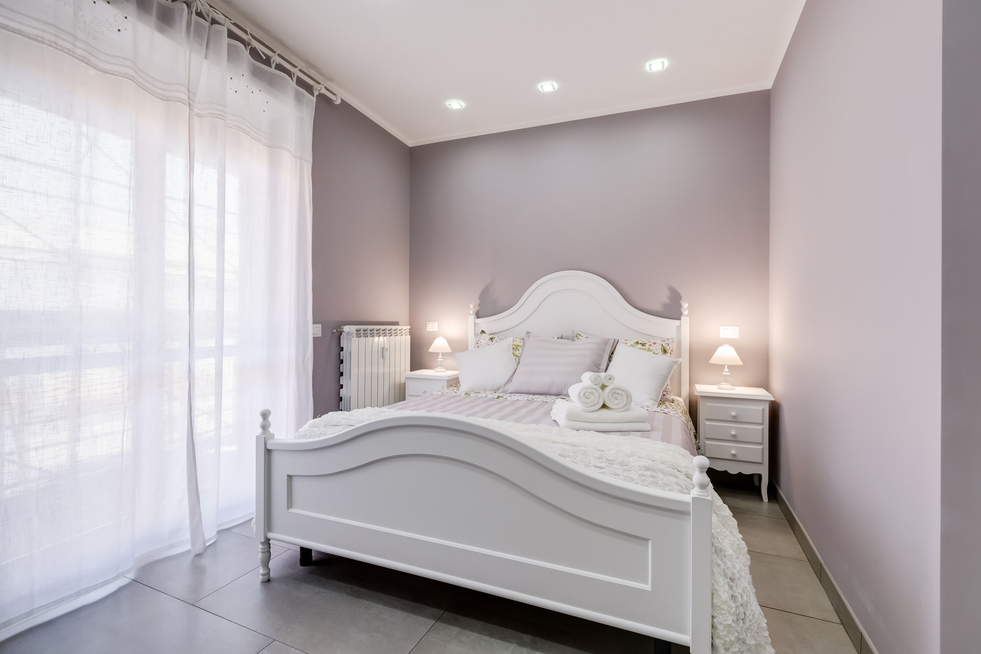 Une chambre avec un lit blanc, deux tables de chevet avec des lampes, un radiateur et des rideaux légers.
