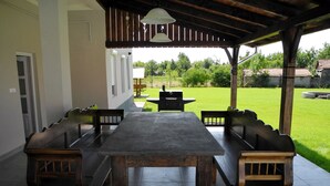 An outdoor patio with a wooden table and benches, a grill, and a view of a green lawn and trees. - Family-friendly holiday home with heated swimming pool for 11 people + 2 baby's (Poroszló)