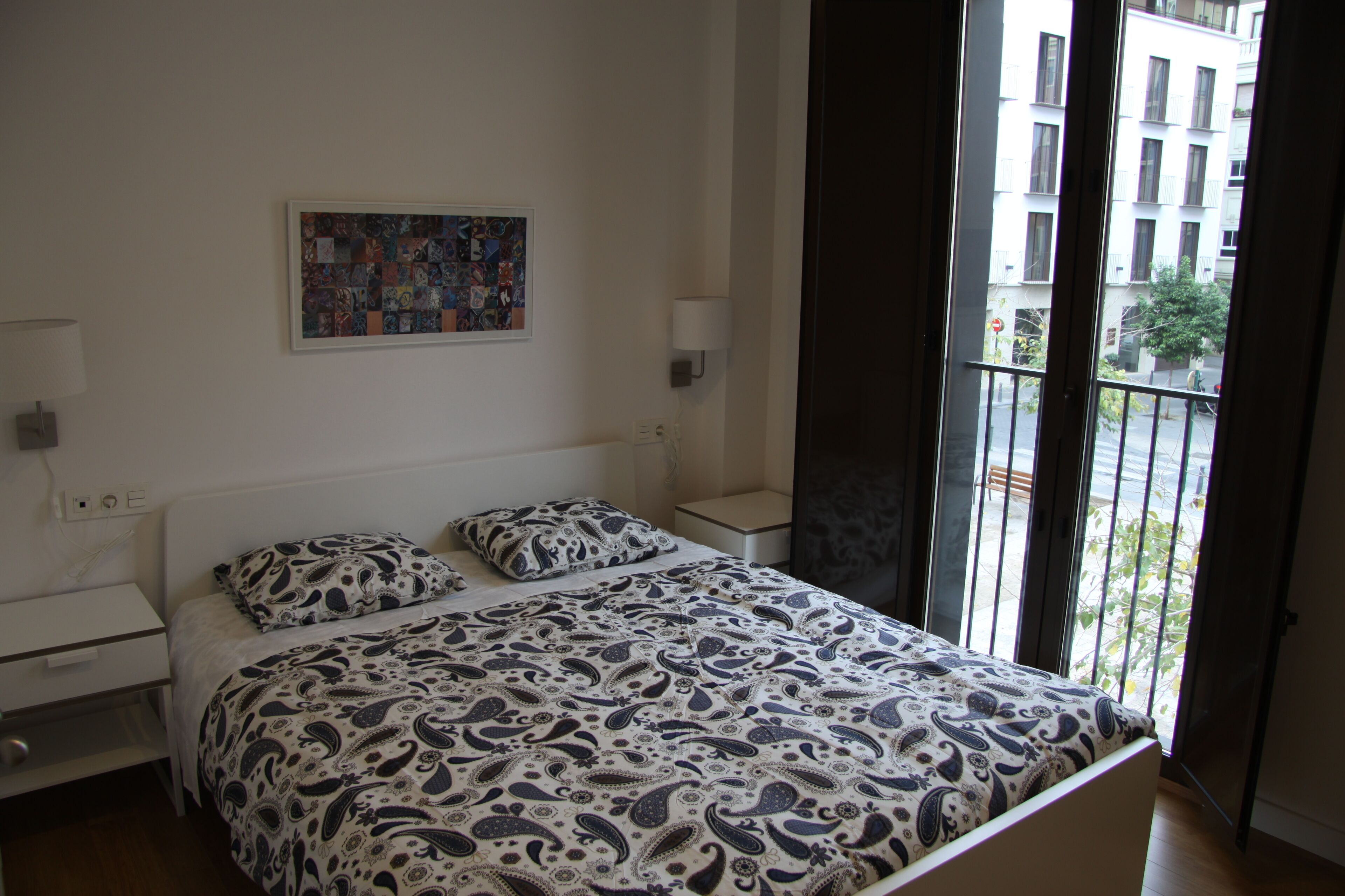 A bedroom with a bed, a nightstand, a lamp, a wall art piece, and a view of the outside through a balcony door.