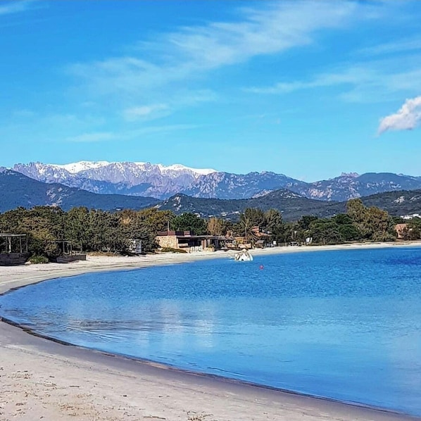 Una playa con mar azul cristalino, orilla arenosa y montañas al fondo.