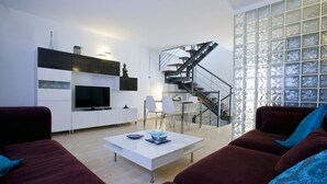 Een moderne woonkamer met een flatscreen televisie, een witte salontafel en een trap naar een balkon.
